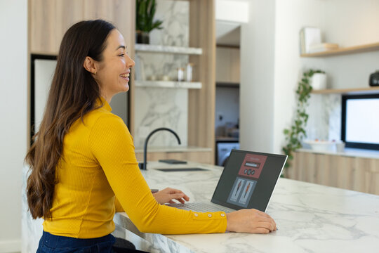 Asian woman sitting at kitchen marble island wearing mustard-yellow ribbed top using laptop