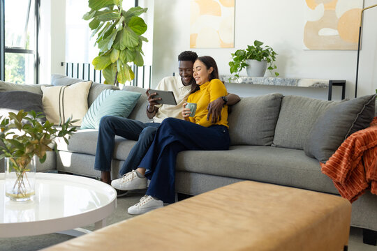 Diverse couple sitting on gray sectional sofa at home, holding tablet and ceramic mug
