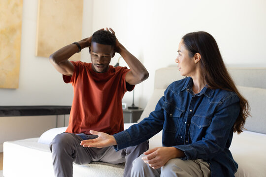 Couple sitting on bed in softly lit bedroom, man holding head with wristband, woman gesturing