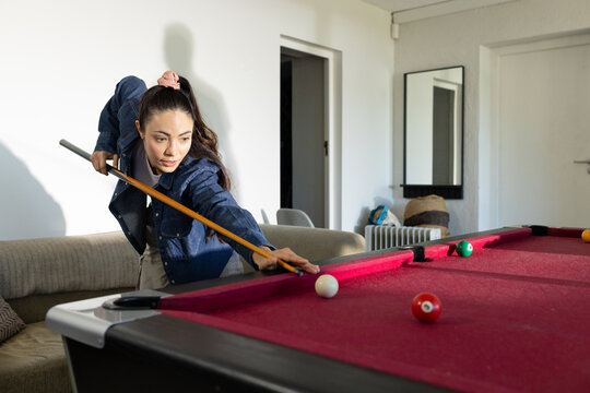 Adult female leaning on red pool-table aiming cue-ball with cue, in denim-jacket at home