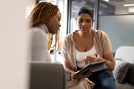 Indian and African American female colleagues reviewing tablet while seated on grey sofa in office