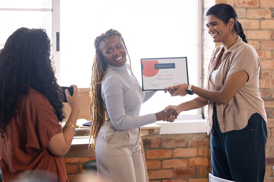 Diverse female team wearing business casual handing certificate, shaking hands in office smartphone