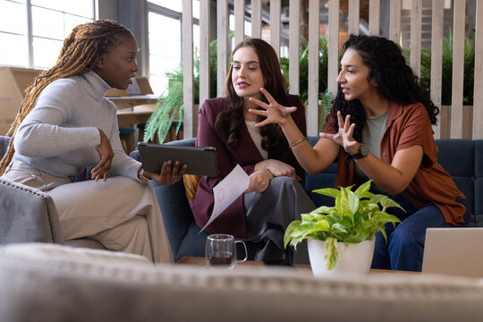 Leaning in, diverse female colleagues in workwear sharing tablet and printed sheet at office