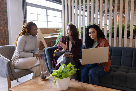 Diverse female team in blazers discussing strategy while reviewing laptop and charts in office nook