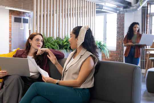 Female coworkers sitting on gray sofa discussing documents and laptop in open plan office lounge