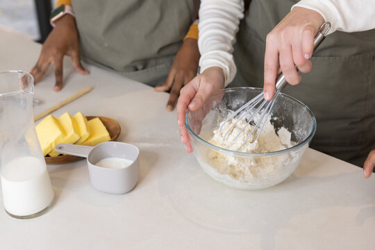 Hands are using metal whisk to mix dough in glass bowl on light countertop with butter
