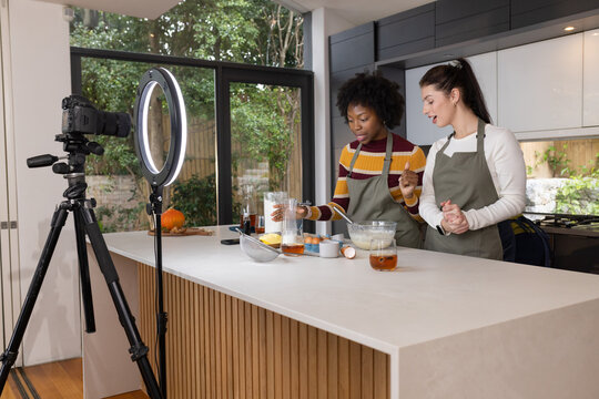 Diverse female hosts preparing batter wearing aprons and filming with DSLR, ring light in kitchen