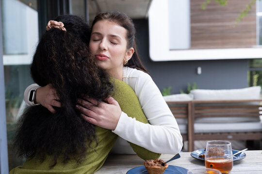 Female friends hugging and leaning into embrace at covered patio table with blue plates and pastry