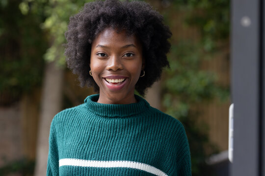 African woman standing and smiling at doorway to garden wearing green sweater and gold hoops