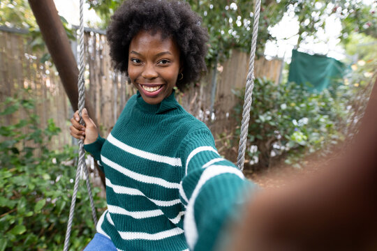 Woman sitting on rope swing in fenced garden wearing green sweater and jeans taking selfie