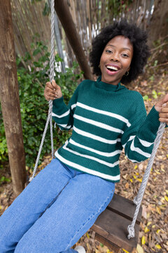 African woman sitting on wooden swing holding ropes in yard wearing green sweater and hoop earrings