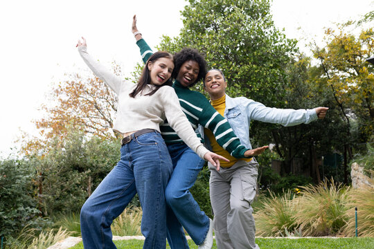 Diverse female friends posing playfully on grassy lawn in park-like garden wearing sweaters, jeans