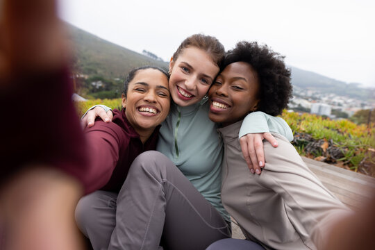 Female friends leaning taking selfie on wood bench on hillside, wearing muted jackets holding phone