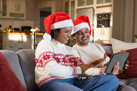 African American couple sitting on gray couch wearing Santa hats, sharing tablet with green ribbon