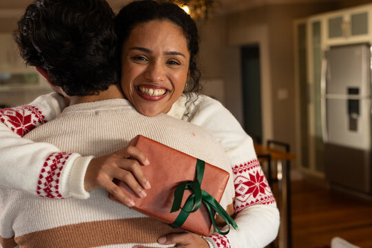 Couple hugging in home kitchen and dining, woman holding wrapped gift, wearing white snowflake knit