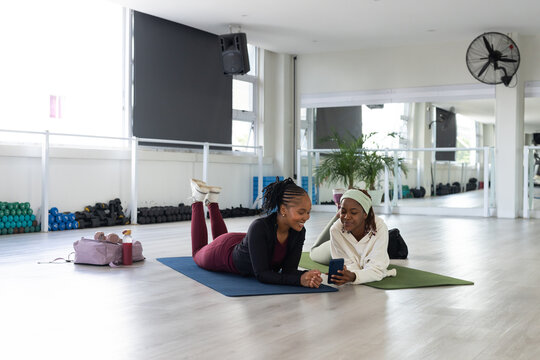 Two African women lying on yoga mats in fitness studio, wearing burgundy leggings, checking phone