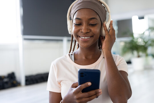 African woman in light tee adjusting headphones and checking blue phone in studio near windows