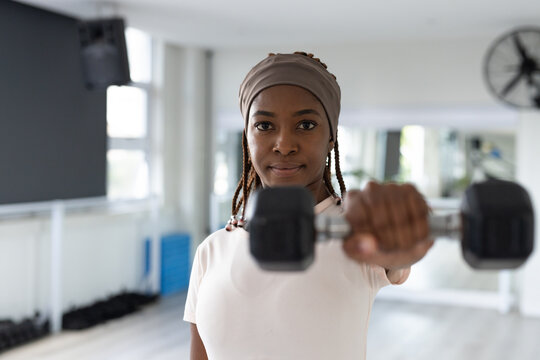 Woman standing and holding hexagonal dumbbell toward camera in fitness studio, wearing headscarf