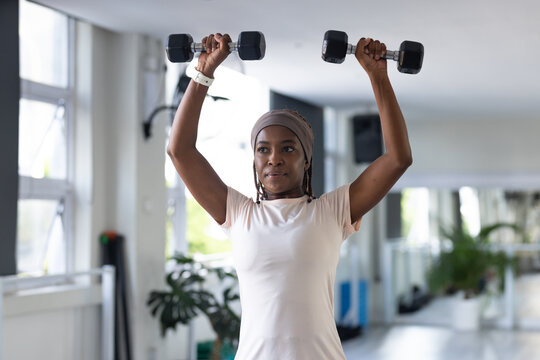 African woman lifting black hex dumbbells overhead in gym, wearing headwrap and wrist tracker