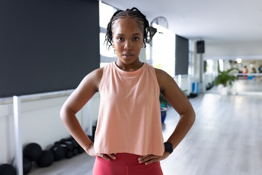 African woman standing hands on hips in gym wearing pink top red leggings near medicine balls