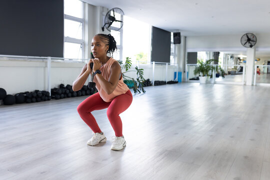 Adult African woman performing goblet squat in gym in white shoes and holding kettlebell near chest