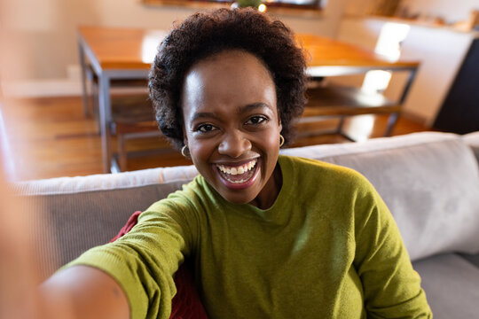 African American woman sitting on couch in living room wearing green top hoops taking phone selfie