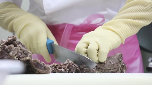 노란 장갑을 낀 두 손으로 칼을 이용해 잘 삶아진 수육 고기를 정성껏 써는 조리 과정 클로즈업 뷰