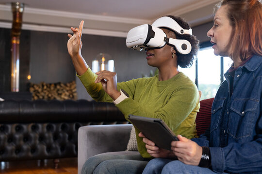 Diverse women wearing green sweater and denim jacket using VR headset and tablet in living room