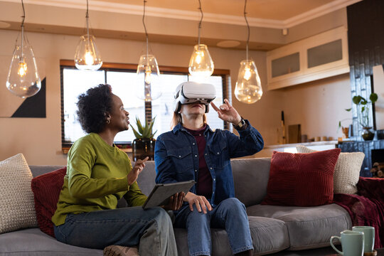 Diverse female friends sitting on sofa at home, right wearing white VR headset, left holding tablet