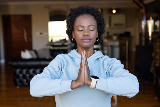 Adult African woman standing pressing palms together at chest in blue hoodie with smartwatch, sofa