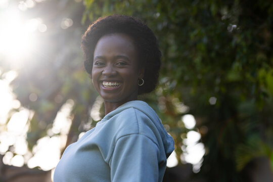 Female smiling and standing in garden wearing light blue hoodie, hoops, curly hair sun flaring