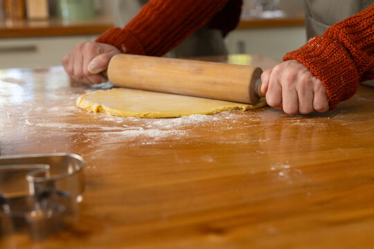 Mature woman rolling dough on floured counter at home with wooden pin, metal cookie cutters, apron