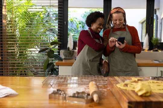 Diverse female friends in green aprons leaning in kitchen at counter checking phone