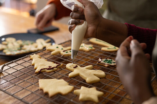 Two female friends decorating human-shaped cookie on wooden table, squeezing piping bag