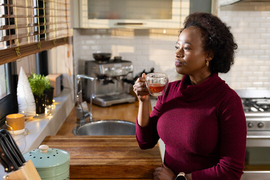 African American woman in burgundy-sweater holding glass cup at counter, gazing out window