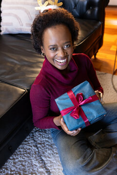 African woman sitting on rug in living room, wearing maroon turtleneck and jeans, holding blue gift