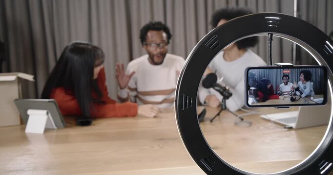 Diverse hosts adjusting to ring light framing phone placing glasses podcasting at wooden table
