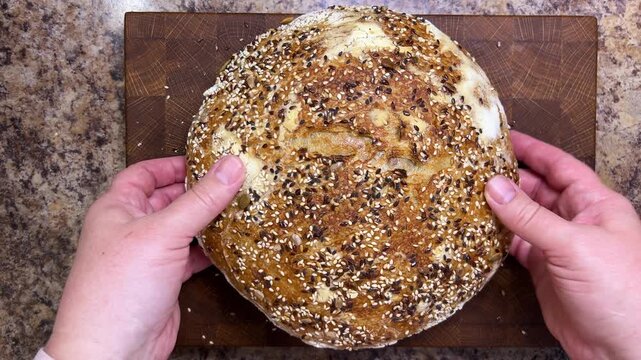 A close-up of female hands holding a round artisan sourdough loaf covered with crunchy sesame and flax seeds, gently pressing the golden crust to show its texture.