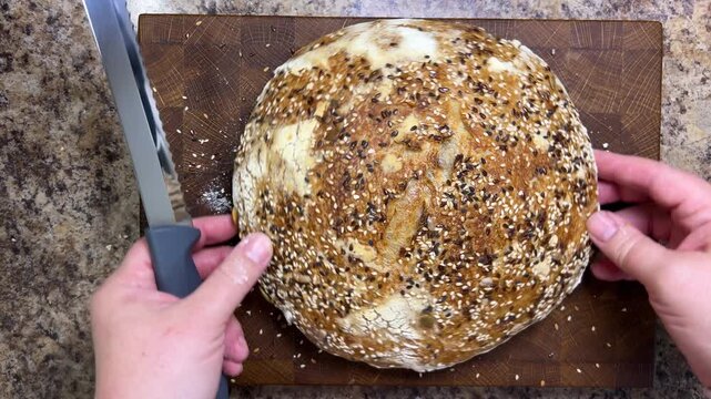 A close-up of a woman slicing a round, seeded sourdough loaf covered in sesame and flax seeds in half on a wooden board with a sharp knife in a kitchen.