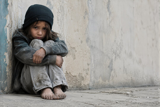 Sad homeless little girl sitting on the cold ground against a dirty street wall in ragged clothes, childhood poverty and despair concept