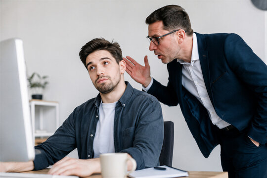 Annoyed young male employee rolling his eyes while his demanding boss is nagging and micromanaging him at the office desk, toxic workplace concept