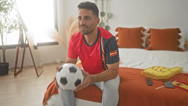 Man on bed holding soccer ball with arms raised cheering in a bedroom building; team pride excitement.