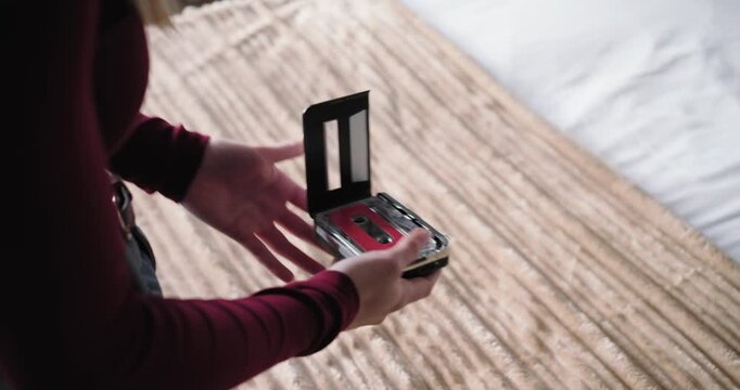 Adult woman maroon top preparing cassette and player with earbuds on bedspread to listen