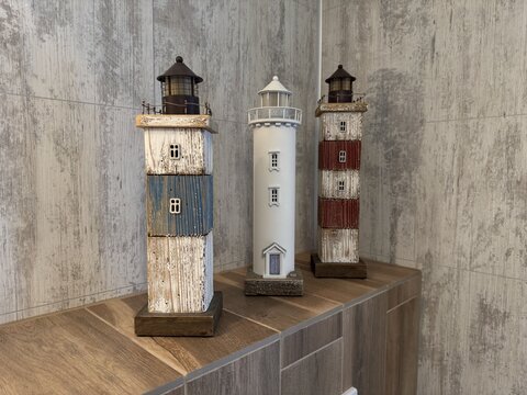 A cozy interior composition featuring three varied decorative lighthouses&mdash;rustic blue, white ceramic, and red striped&mdash;standing in a row on a wooden shelf against grey textured wall panels.