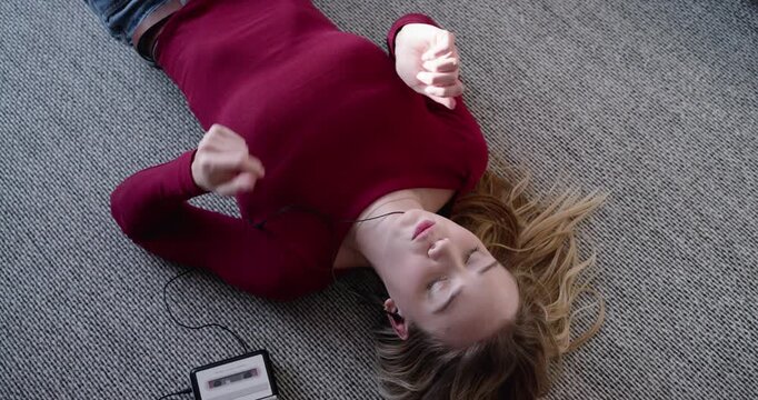 Female lying on textured carpet, listening to cassette through earbuds, mouthing and tapping