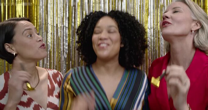 Diverse female friends celebrating after center woman clapping, blowing gold blowers at photobooth