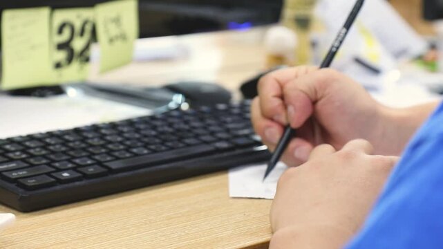 책상 위 키보드 옆에서 펜을 쥐고 메모지에 무언가를 적는 인물의 손 클로즈업 뷰