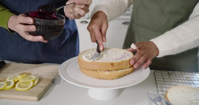 Diverse women in aprons smoothing frosting spooning compote and stacking cake on stand assembling