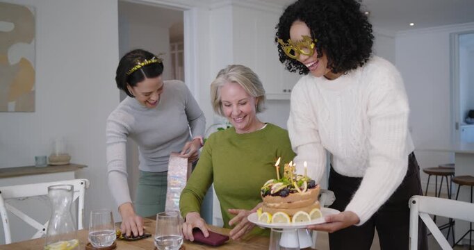 Women friends marking birthday in kitchen, friend with star glasses carrying cake, sashing