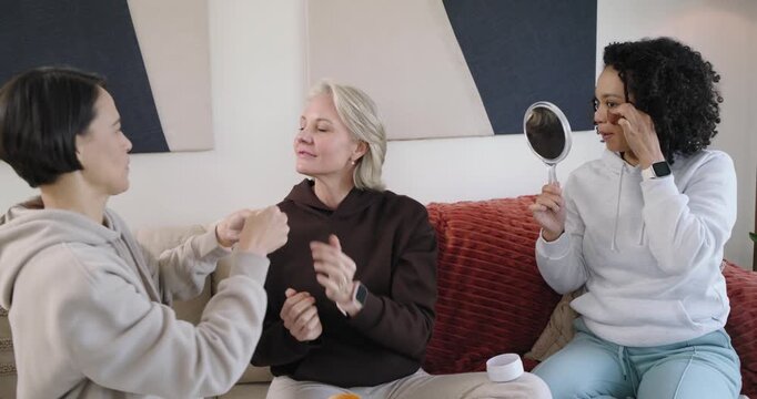 Diverse women friends left friend picking under-eye pink patch applying on couch